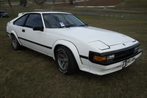 Biała Toyota Supra MK2 z lat 80. stoi na trawie, z widokiem na tor wyścigowy w tle.