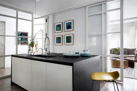 Isla de cocina moderna con encimera oscura, separada del salón por una pared de cristal.