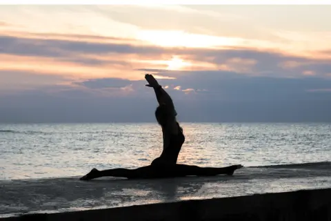 Silhouette di una donna in Ardha Hanumanasana sulla riva del mare al tramonto, con le braccia alzate verso il cielo.
