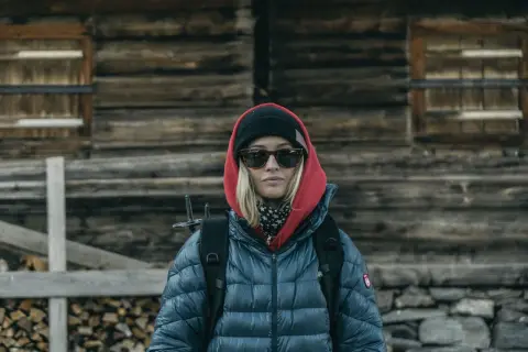 Kobieta w czerwonej bluzie z kapturem i czarnej czapce, w okularach przeciwsłonecznych, z plecakiem, przed drewnianą chatą.