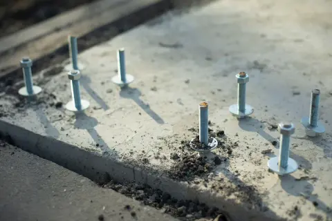 Kilka metalowych kotew z podkładkami i nakrętkami osadzonych w betonie.
