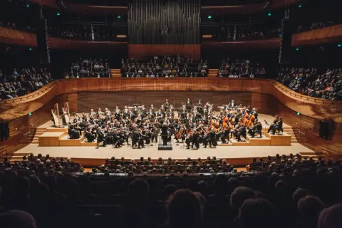 Orkiestra symfoniczna grająca na scenie w sali koncertowej, przed publicznością.