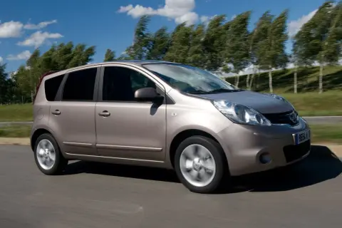 Złoty Nissan Note 2010 pędzi po drodze, drzewa na tle falują na wietrze.