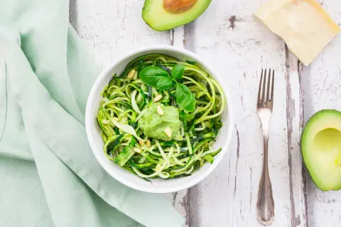 Zoodles con pesto di avocado, guarniti con pinoli e basilico. Accanto, mezzo avocado, un pezzo di parmigiano e una forchetta.