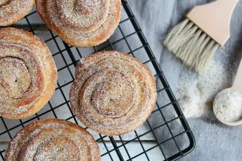 Nybakade kanelbullar med strössel av brunt socker och pärlsocker på ett svart galler. En träpensel och en sked med socker ligger bredvid.