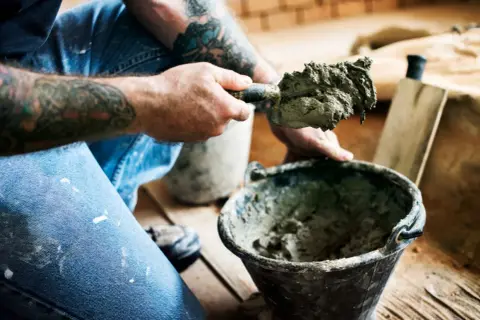 Ręce z tatuażami nabierają zaprawę kielnią z wiadra.