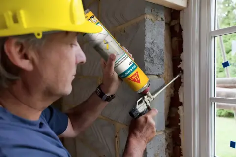 Hombre con casco amarillo usa sellador de poliuretano para rellenar huecos en instalación de ventana.