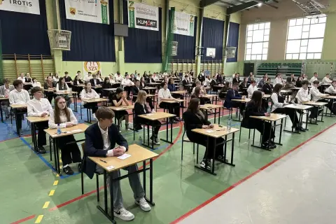 Uczniowie siedzą przy ławkach w sali gimnastycznej, czekając na wyniki egzaminu ósmoklasisty.