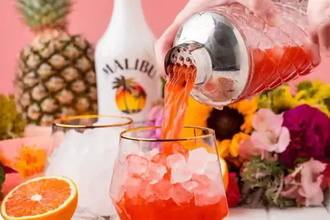 Orzeźwiający drink z malibu, nalewany z shakera do szklanki z lodem. W tle ananas, butelka Malibu i kwiaty.