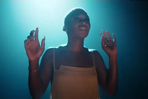 Cynthia Erivo stand up, wznosi ręce w górę, w świetle reflektorów.