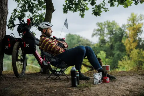 Mężczyzna odpoczywa podczas wycieczki rowerowej, gotując posiłek na kuchence turystycznej. Obok rower z sakwami.