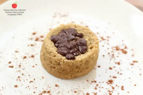 Delicioso mug cake saludable con trozos de chocolate negro, espolvoreado con cacao en polvo sobre un plato blanco.