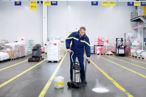 Hombre realiza limpieza almacen con pulverizador. A su lado, un bidón de líquido y una fregadora industrial.