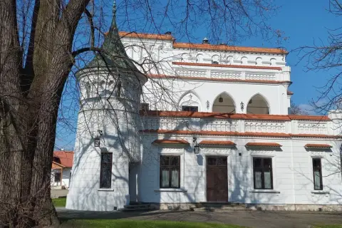Biały pałac z wieżyczką i czerwonym dachem, otoczony drzewami. W oddali widać inne budynki. To piękny widok na **Radomyśl Wielki**.