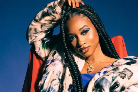 Nija Charles, with striking blue eye makeup and braids, poses confidently in a colorful jacket against a blue backdrop.