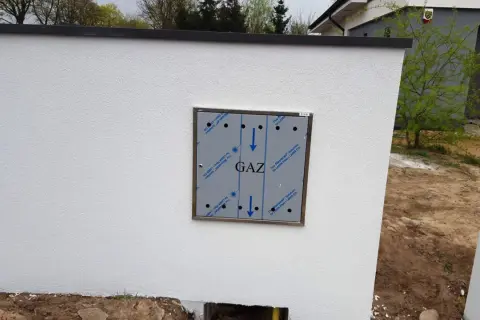 Metalowa skrzynka gazowa z napisem "GAZ" zamontowana na białym, otynkowanym murze.