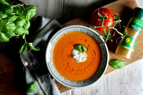 En skål med krämig tomatsoppa, gjord på krossade tomater, toppad med en klick gräddfil och basilika. En färsk tomat och en flaska Knorr Fresh Vegetable Vegan Fond ligger bredvid.