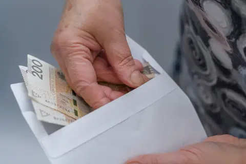 Dłoń wkłada banknoty 200 zł do koperty. Ilustracja pokazuje przykłady przeliczenia emerytury kapitałowej na emeryturę powszechną.