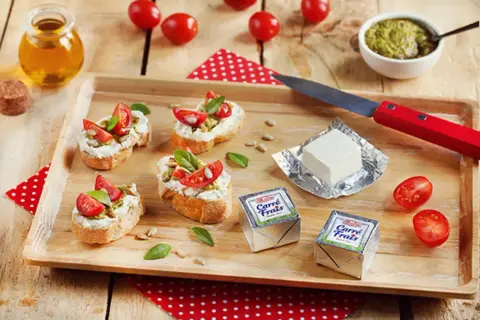 Bruschettas garnies de fromage frais, tomates et pesto, avec des emballages de Carré Frais le cendre et des tomates cerises sur un plateau en bois.