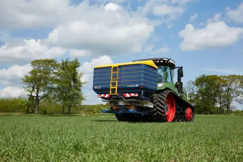 Zielony traktor z przyczepą rozsiewającą na polu. Na przyczepie widoczna nazwa "bogballe tabela wysiewu".
