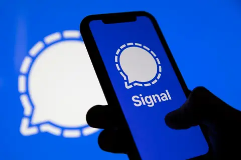 Dłoń trzyma smartfon z otwartą aplikacją Signal. W tle widoczne logo Signal.