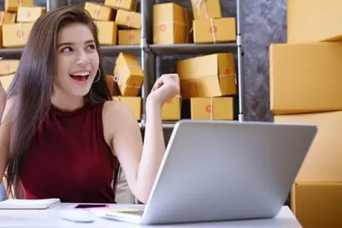 Szczęśliwa młoda kobieta przy laptopie, otoczona paczkami. To inspiracja, jak zacząć dropshipping i rozwijać swój biznes online.