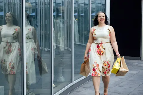 Uśmiechnięta kobieta w kwiecistej sukience plus size, z torbami zakupowymi w rękach, idzie obok witryny sklepowej.