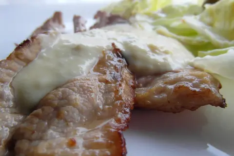 Tiernas lonchas de blanqueta de cerdo cubiertas con una cremosa salsa blanca, acompañadas de hojas verdes frescas.