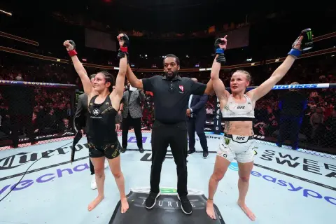 Two female fighters stand with arms raised, a referee between them. The outcome of their boxing draw is announced to the roaring crowd.