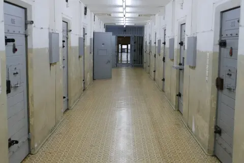 Korytarz więzienny z rzędami cel. Drzwi cel, niektóre z numerami jak 106 i 222, są zamknięte. W głębi widać kraty i otwarte drzwi.
