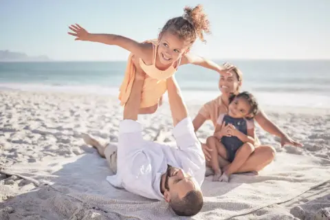 Rodzina na plaży na Malcie. Ojciec podrzuca córkę, matka z drugą córką siedzą obok. Czyste niebo, spokojne morze, piasek. Malta bezpieczeństwo.