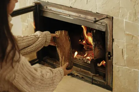 Une personne en pull ajoute une bûCHE dans un poêle à bois qui fume, porte ouverte. La chaleur des flammes réchauffe la pièce.