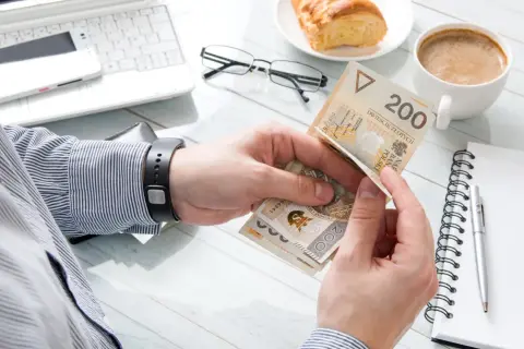 Ręce liczą polskie banknoty 200 zł. Obok laptop, kawa, rogalik i notatnik. To obrazek pokazujący klasa średnia zarobki.