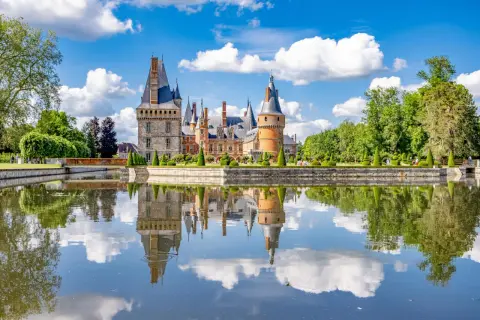 Magnifique château en Eure-et-Loir, reflet parfait dans l'eau calme, entouré de jardins à la française et d'arbres verdoyants sous un ciel bleu parsemé de nuages.