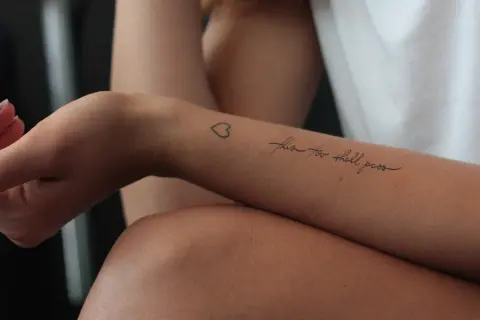 Ein Tattoo auf dem Unterarm einer Frau mit der Aufschrift "this too shall pass" und einem kleinen Herz. Ein Symbol der Hoffnung nach der Überwindung von Alkoholsucht.