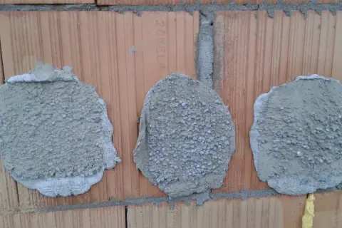 Trzy plamy zaprawy z widocznymi w nich kołkami na tle cegły.