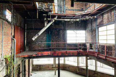 Intérieur d'une usine abandonnée, typique d'un site d'urbex dans la Somme. Structures métalliques rouillées, tuyaux, briques apparentes et lumière filtrant par les fenêtres.