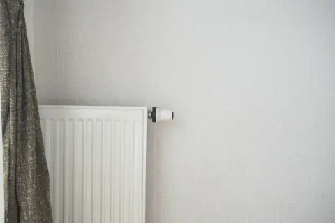 Radiateur blanc moderne, prêt à être alimenté par une pompe à chaleur. Un rideau discret complète la scène.
