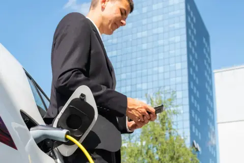 Uomo in giacca e cravatta controlla il telefono mentre la sua auto elettrica si ricarica, pianificare viaggio auto elettrica è facile con la tecnologia.
