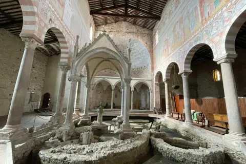 Interno della Basilica di San Piero a Grado, con affreschi medievali e resti archeologici al centro.