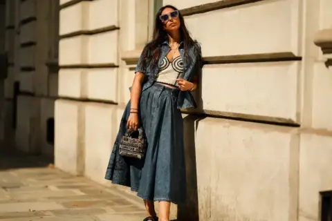 Mujer con gafas de sol posa apoyada en una pared, luciendo un conjunto vaquero y una falda vaquera larga.