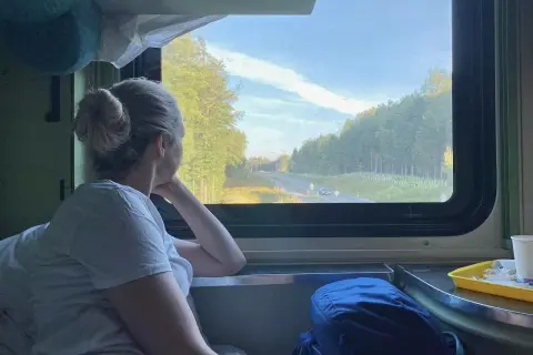 Frau blickt aus dem Fenster eines Nachtzuges nach Italien. Die Landschaft zieht vorbei, Bäume und eine Straße sind zu sehen.