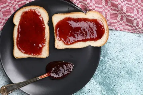 Zwei Scheiben Toast mit leckerer Jostabeeremarmelade und ein Löffel voll davon auf einem schwarzen Teller.