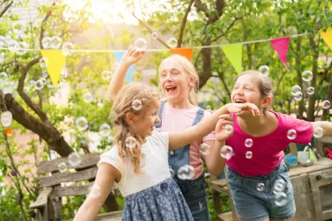 Trzy dziewczynki śmieją się i łapią bańki mydlane w ogrodzie. Wesołe życzenia urodzinowe dla dziecka!