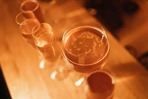 Kilka kieliszków z bursztynowym napojem, jeden z nich z widocznym napisem "kombucha".