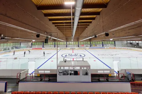 Eishalle Wiehl mit Eisfläche, Banden und Tribüne. Einige Eisläufer sind auf dem Eis zu sehen.
