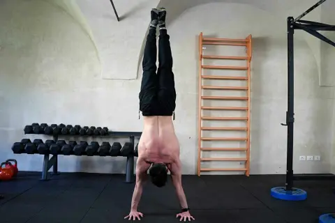 Uomo esegue flessioni per spalle in un centro fitness, con bilancieri e spalliera svedese sullo sfondo.