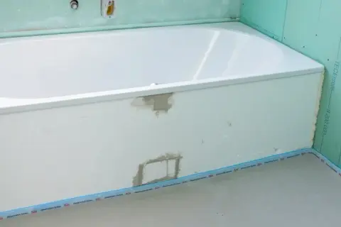 Einbau einer neuen, weißen Badewanne in einem Badezimmer im Rohbau. Grüne Gipskartonplatten und vorbereitete Rohre sind sichtbar.
