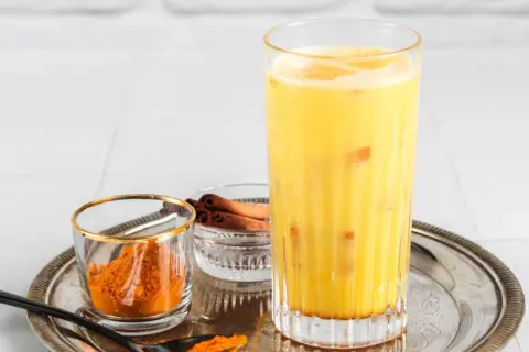 Ein Glas goldene Milch mit Kurkumapulver und Zimtstangen daneben.