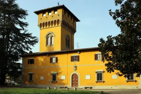 Edificio giallo con torre merlata a Borgo San Lorenzo, circondato da alberi verdi e un cielo azzurro.